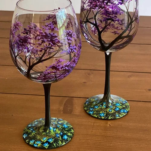 Hand hält handbemaltes Weinglas mit Jahreszeiten-Baum-Motiv, betont die Eleganz des Kunstglases & Weinliebe.